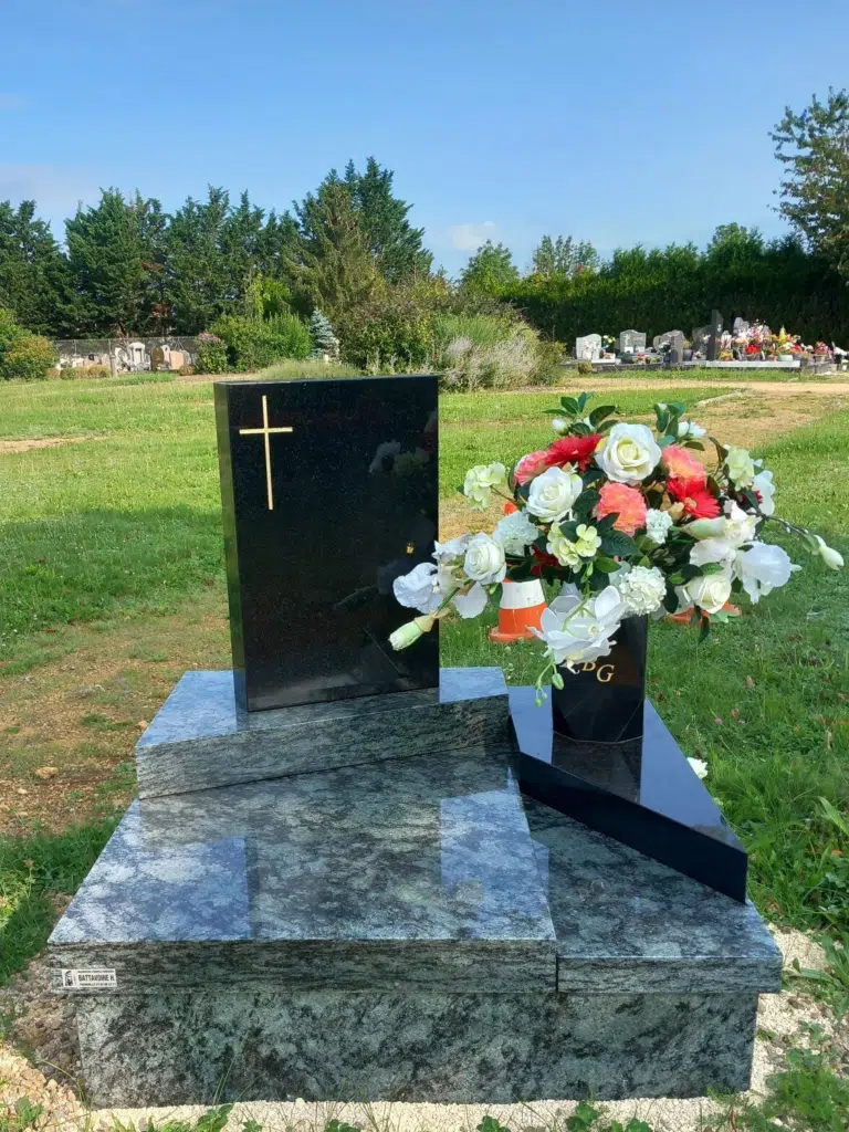 Petit monument cinéraire noir avec stèle dorée et bouquet de fleurs.